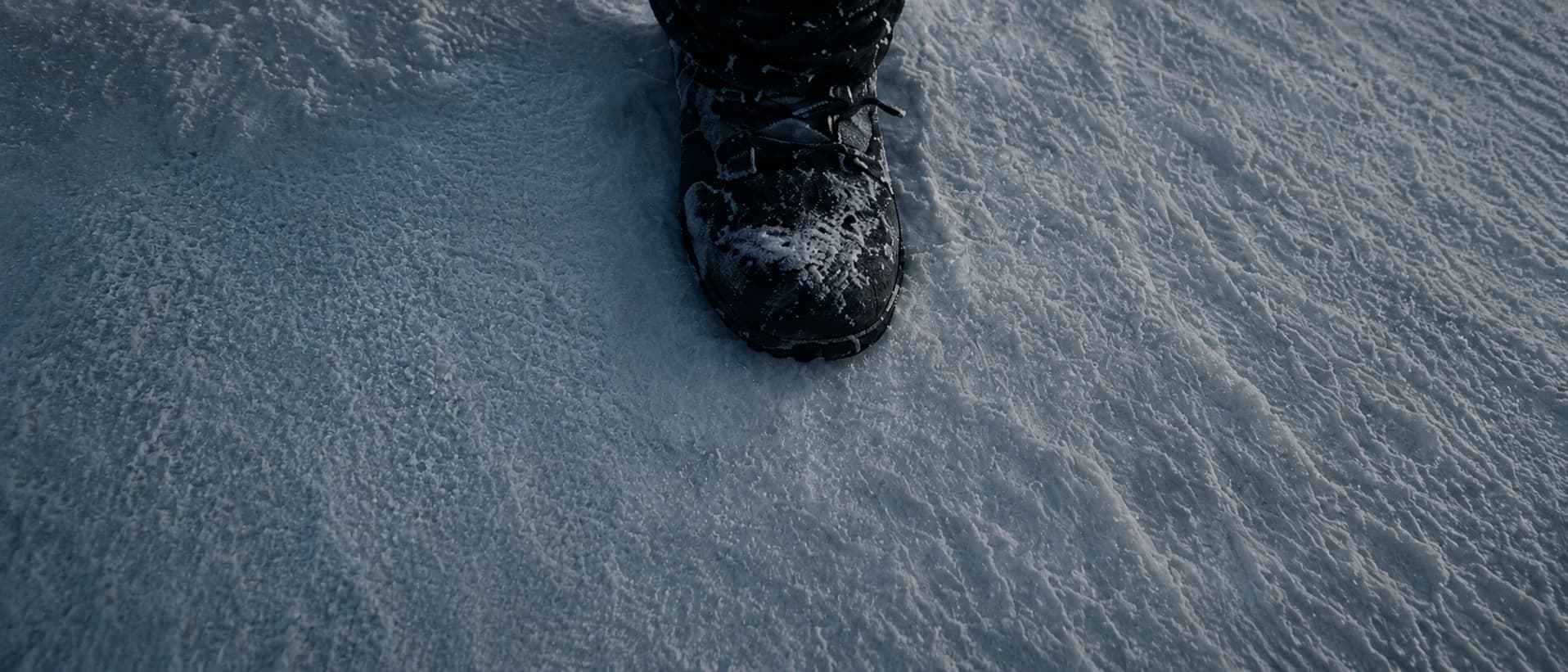 Storyboard frame — top-down high angle of a single boot pressing into wind-rippled snow.